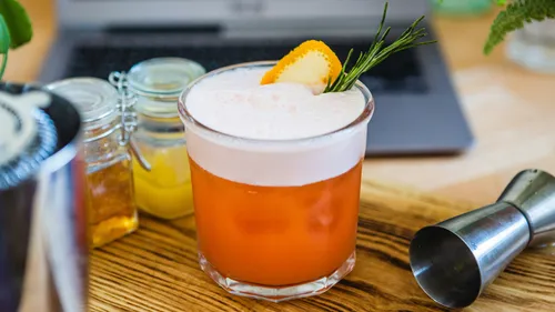 Cocktail im Glas mit Schaum, auf Holztisch neben Laptop.
