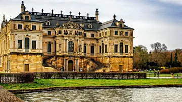 Historic building stands on the water, people walk next to it.