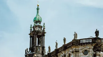 Kirchturm mit grünem Dach und statuenumrandetem Balkon.