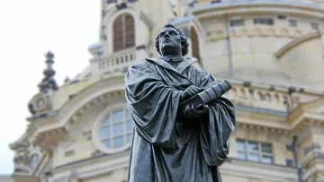 Bronzestatue eines Mannes mit Buch vor einem historischen Gebäude.
