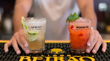 Zwei Cocktails stehen auf einer Theke, Hände halten sie.