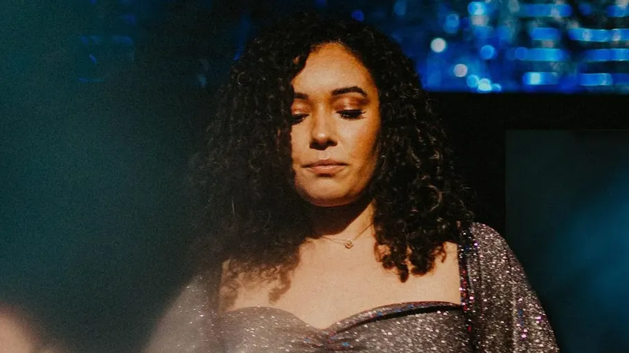 A woman with curly hair stands thoughtfully on a stage.