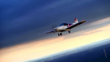 An airplane flies through cloudy skies over a landscape.