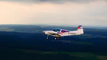 An airplane flies over a green landscape under clouds.