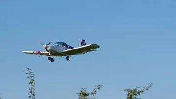 An airplane flies over trees under clear skies.
