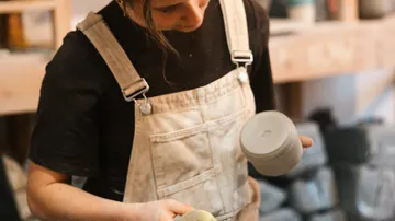 A woman sculpts a gray clay cup in a workshop.