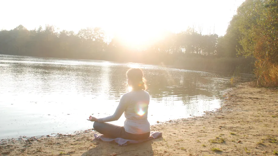 A person meditates on a calm lake in sunlight.