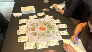 Game board with cards, people discussing and writing notes.