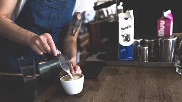 Barista gießt Milch in eine Tasse Kaffee an einer Theke.