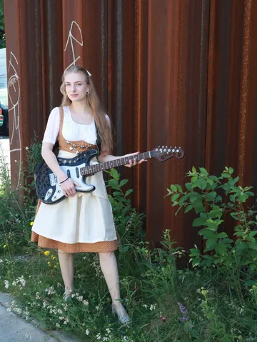 Frau steht mit Gitarre vor rostigem Metallhintergrund.