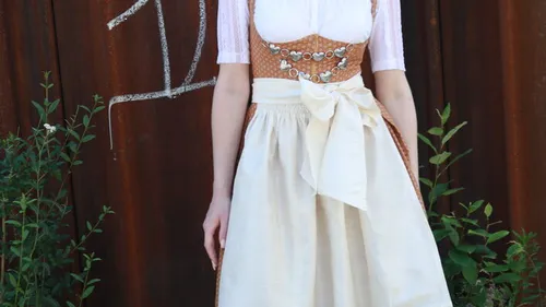 Frau im Dirndl steht vor rostiger Wand, umgeben von Blumen.