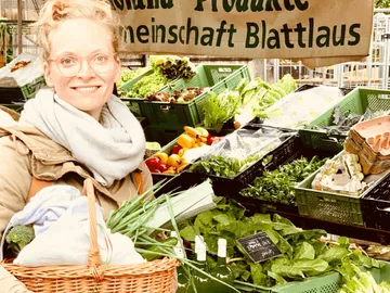 Frau mit Korb lächelt vor einem Marktstand voll Gemüse.