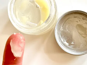 Finger mit Creme, die aus einem Glasbehälter entnommen wird.