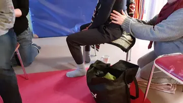 Person sitzt auf Stuhl, während andere unterstützen; bunte Umgebung.