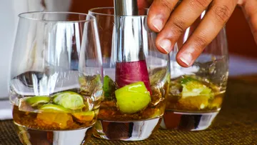 Ein Glas wird mit einem Stößel gemixt, Limettenstückchen enthalten.