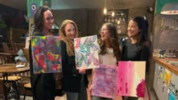 Four women hold colorful paintings in a bar.