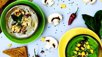 Two bowls of soup with herbs, surrounded by ingredients and bread.