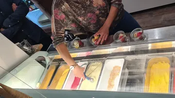 Ice cream saleswoman scoops ice cream from a refrigerated glass counter, customers wait.