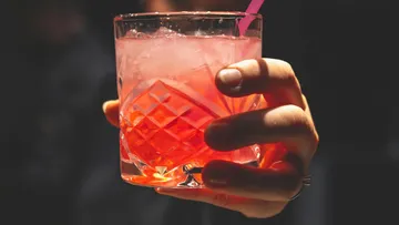 A glass with a pink cocktail is held, the background is dark.