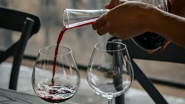 A hand pours red wine into a glass on a table.