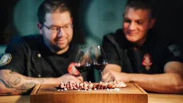 Two chefs enjoy artistically prepared meat and wine.
