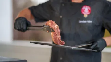 Cook holds a piece of meat over a grill with tongs.