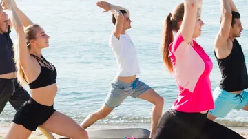 Fünf Personen machen Yoga am Strand, Wasser im Hintergrund.