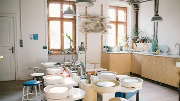 Keramikwerkstatt mit Töpferbänken und Pflanzen im Hintergrund.