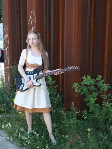 Junge Frau im Dirndl spielt E-Gitarre vor rostigem Hintergrund.