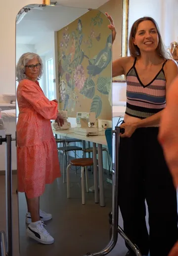 Two women pose in front of a mirror in a bright room.