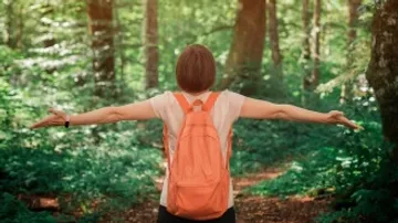 Eine Person mit Rucksack steht im Wald und breitet Arme aus.