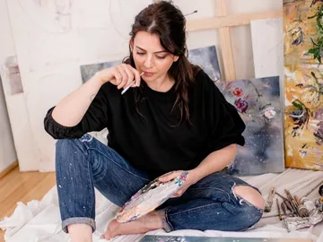 Junge Frau sitzt im Atelier und malt konzentriert auf Leinwand.