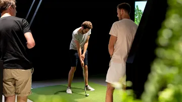 Man putts golf ball in an indoor golf environment, spectators watch.