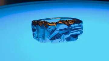 Ein Ring mit detaillierter Skulptur leuchtet auf blauem Hintergrund.