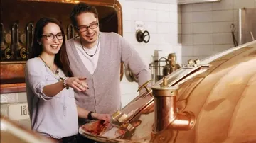 Two people watch a copper kettle in a brewery.