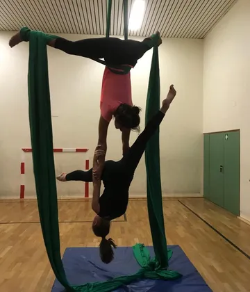 Two women are acrobatically hanging from green cloths in a room.