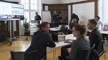 Gruppendiskussion, Präsentation, helles Raum, Bildschirme sichtbar.