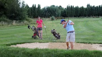 Mann schlägt Golfball aus Bunker; Frau beobachtet in der Nähe.