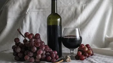 Red wine bottle is on the table next to glass and grapes.