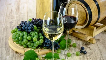 Glasses of wine stand next to grapes and a wooden barrel.