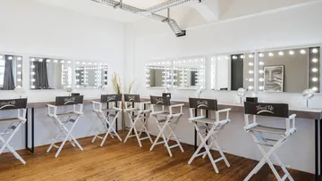 Barber chairs stand in front of mirrors with theatrical lights in a room.