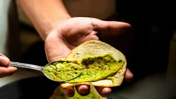 Hand holds tortilla while spoonfuls of guacamole are spread over it.