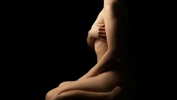 Naked person kneels, holds himself with one hand. Dark background.