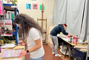 Two women work creatively in a studio full of materials.
