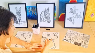 A woman draws floral patterns on paper in front of pictures.