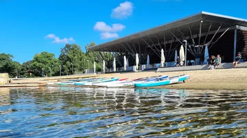 Paddleboards are located on the shore next to a modern building.