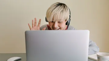 Young person with headphones waves smiling in front of laptop.