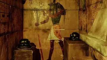 Wandmalerei zeigt Anubis in antikem Grabraum mit Statuen.