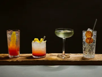 Four cocktails sit on a wooden board against a black background.