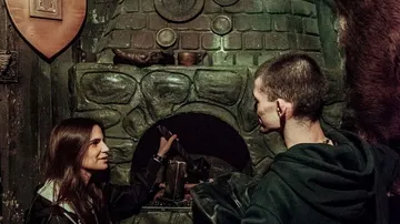 Zwei Personen stehen vor einem steinernen Kamin und sprechen.